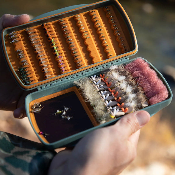 Open fly fishing lure case with various lures held by a person against a blurred natural background.