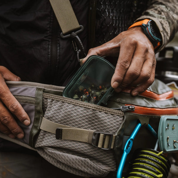Person opening a waist bag with camouflage clothing and outdoor gear in the background