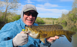 Person holding a large fish by a river with trees and blue sky in the background